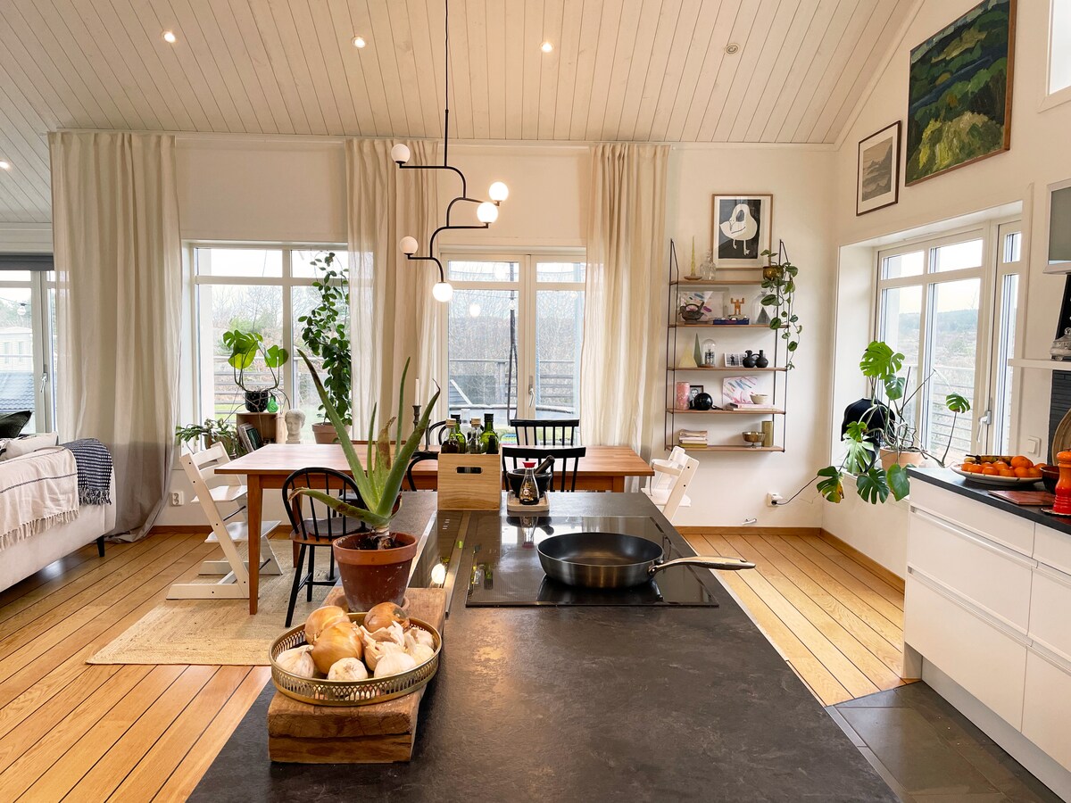 An open-plan kitchen and dining area is featured, illuminated by natural light from large windows. A long dining table with seating is visible, alongside plants that add a touch of greenery. A sleek island with a skillet and a bowl of bread completes this inviting space.