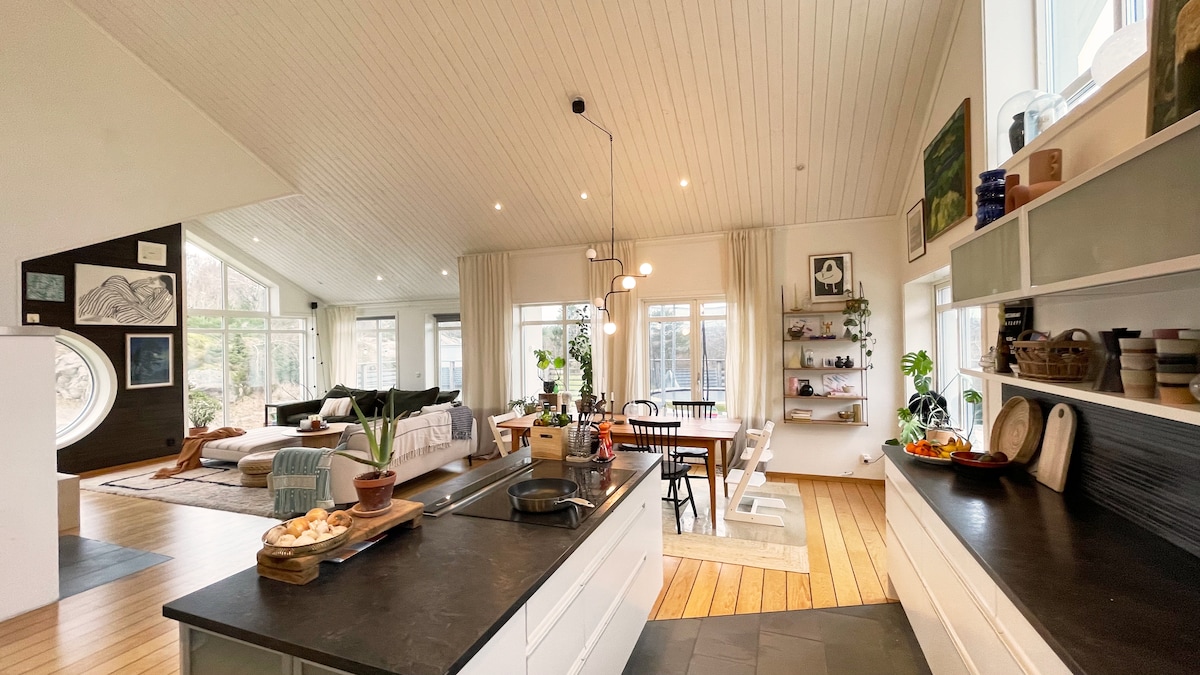 An open-plan kitchen and living area are presented, featuring large windows that allow ample natural light. A spacious island is positioned in the kitchen, surrounded by seating. A dining table with chairs is set against the backdrop of a cozy living space, adorned with plants and artworks.