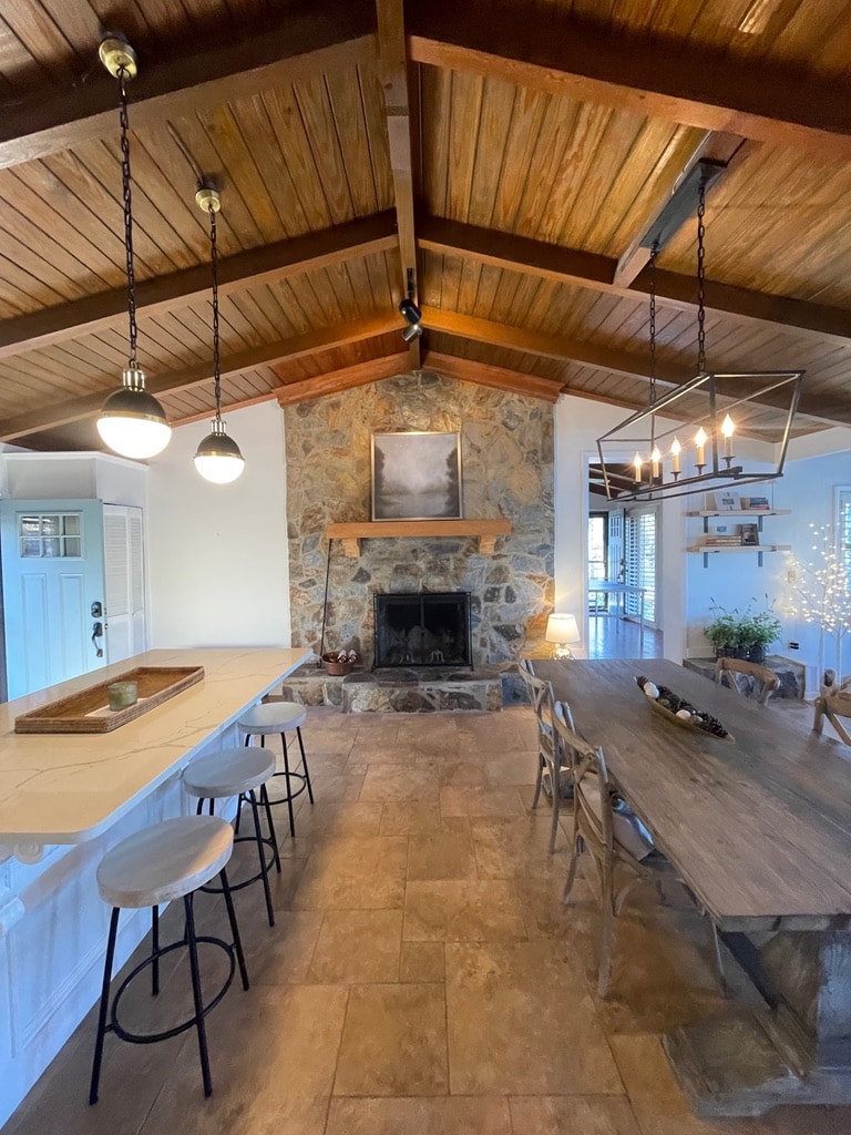 An open concept living area features vaulted wood ceilings with pendant lighting. A stone fireplace is centered against a wall, accompanied by a large framed artwork. A dining table is situated on one side, with a breakfast bar equipped with stools on the opposite side.