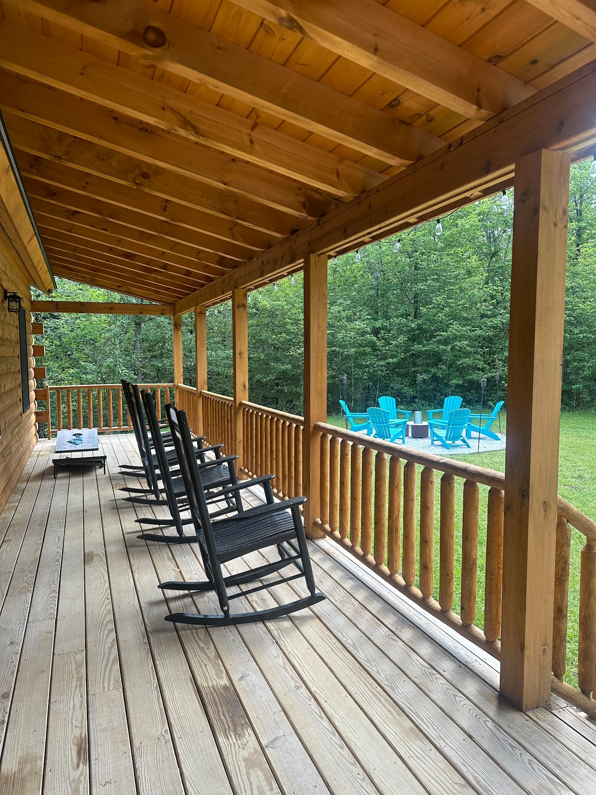 A spacious porch features a series of black rocking chairs arranged along a wooden railing. An outdoor seating area with blue chairs is visible in the yard, surrounded by greenery, inviting moments of relaxation in a tranquil setting.