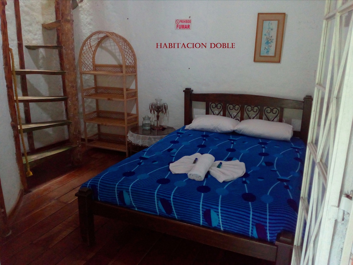 A cozy bedroom features a spacious bed dressed in blue linens, accompanied by two rolled towels. A shelving unit and a framed artwork add to the room's charm. Light wooden floors and a staircase contribute to the overall warm ambiance of the space.
