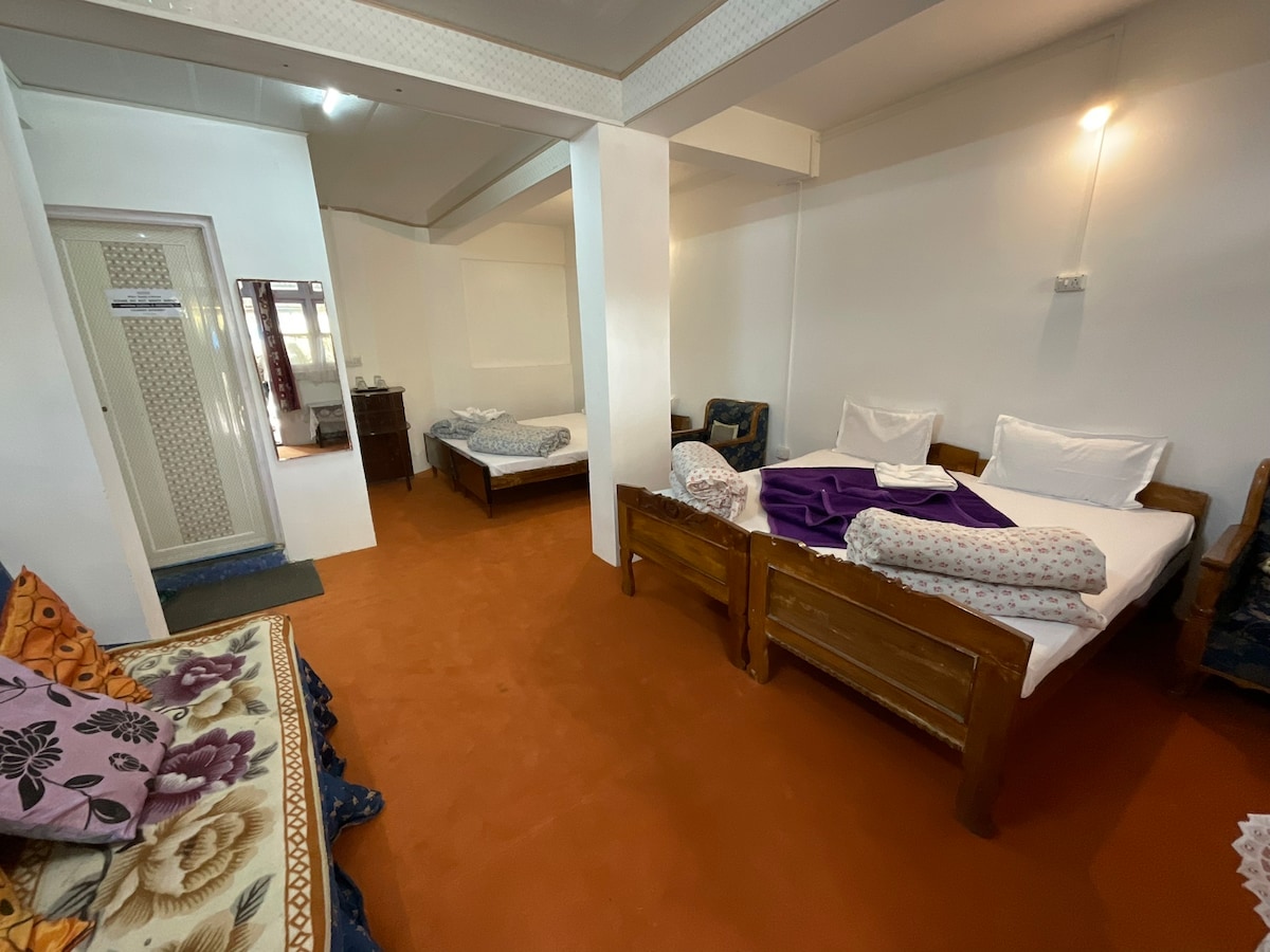 A spacious family room features two wooden beds, each made with simple bedding. A colorful sofa chair and a textile with floral patterns are visible in one corner. Warm orange carpeting covers the floor, enhancing the room's welcoming feel.