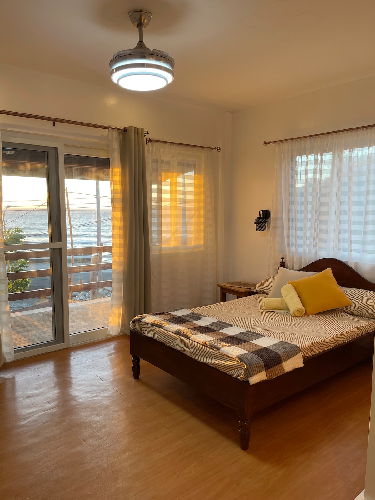 A bright and airy bedroom features a spacious bed adorned with a checkered blanket and a soft yellow pillow. Light filters through sheer curtains, casting a warm glow. A small nightstand is positioned beside the bed, and sliding glass doors open to a scenic view.