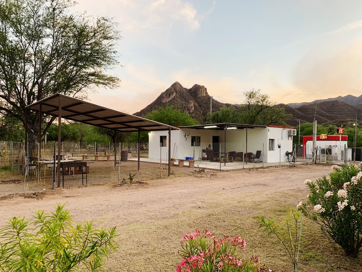 A single-story building is surrounded by a natural landscape, featuring a covered patio area with seating. Nearby, a mountain rises in the background, with a pathway leading through flowers and shrubs that enhance the outdoor setting.