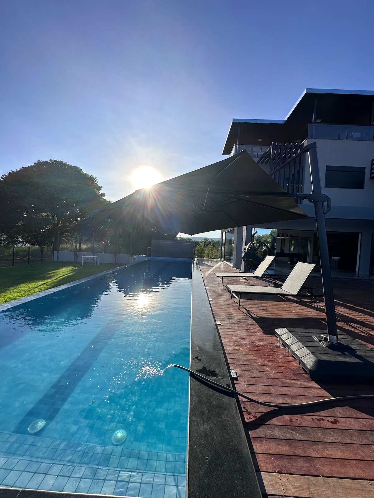 A swimming pool is situated alongside a wooden deck, with sun loungers positioned for relaxation. A large umbrella provides shade over part of the area. The sun is visible in the background, casting light across the pool and surrounding landscape.