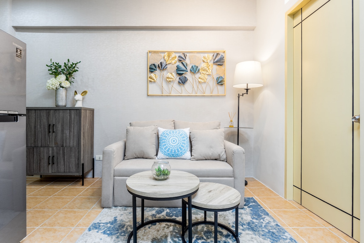A cozy living space features a light-colored sofa with a decorative cushion, complemented by two round coffee tables. A wall art piece showcasing subtle leaf designs is mounted above the sofa. A cabinet and floor lamp complete the warm and inviting atmosphere.