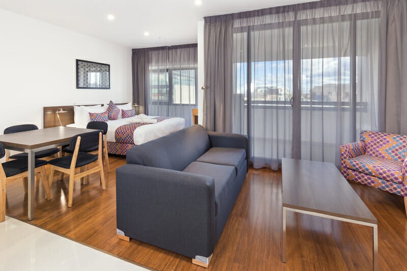 A modern living space is showcased, featuring a dark fabric sofa and a wooden coffee table. A dining area with four chairs is visible, while light filters through large windows dressed in sheer curtains, illuminating a cozy sleeping area with patterned bedding.