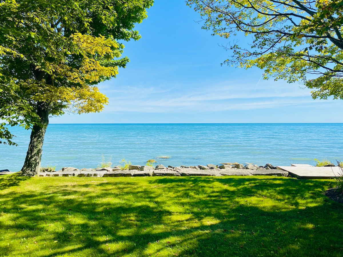 A serene view of Lake Michigan is captured from the grassy yard, framed by leafy trees. The calm water stretches to the horizon, with smooth rocks lining the shore. Soft sunlight enhances the peaceful ambiance of this lakeside setting.