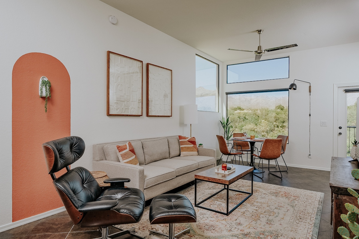 A bright living space showcases a neutral sofa paired with a mid-century black lounge chair. Large windows provide natural light and mountain views. A modern dining table and chairs are positioned near the view, complemented by an accent wall in a warm tone.