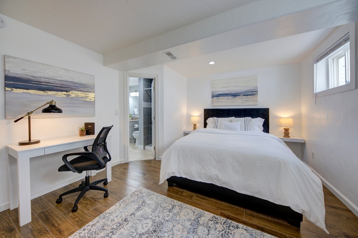 A bright bedroom features a spacious bed with crisp white linens, complemented by two bedside lamps. A desk with a chair is positioned against one wall. Natural light enters through a window, enhancing the modern artwork above the bed and the warm wood flooring.