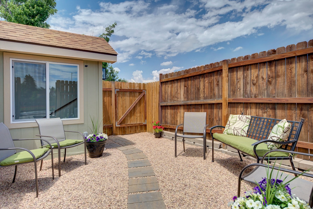 A private courtyard features a stone pathway surrounded by gravel, leading to seating options, including two chairs and a bench, all designed for relaxation. Flower pots add color to the space, with a wooden fence providing privacy and a clear blue sky overhead.