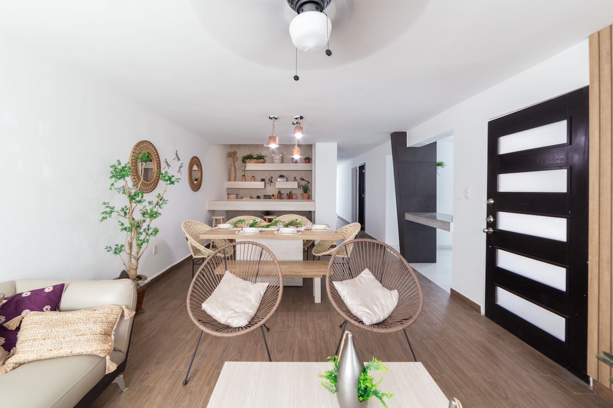 A spacious living area features a neutral color palette with light wood flooring. A modern dining table is set, surrounded by decorative plants. Two comfortable chairs frame a coffee table, while a ceiling fan provides air circulation. A stylish entrance door adds character to the space.
