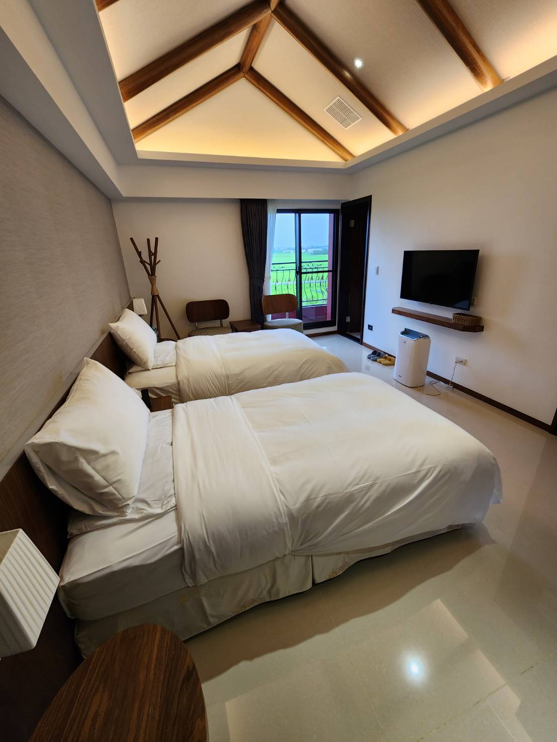 The room features two single beds with crisp white linens, positioned on polished flooring. A television is mounted on the wall opposite the beds. Natural light enters through a large window, and a small seating area is visible beside a minimalist coat rack.