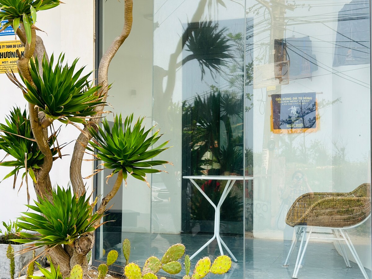 A sleek entrance showcases large glass doors framed by a vibrant green succulent plant. A white table and a stylish chair are arranged outside, inviting relaxation. In the background, palm trees provide a tropical feel, while a framed artwork is visible on the wall.