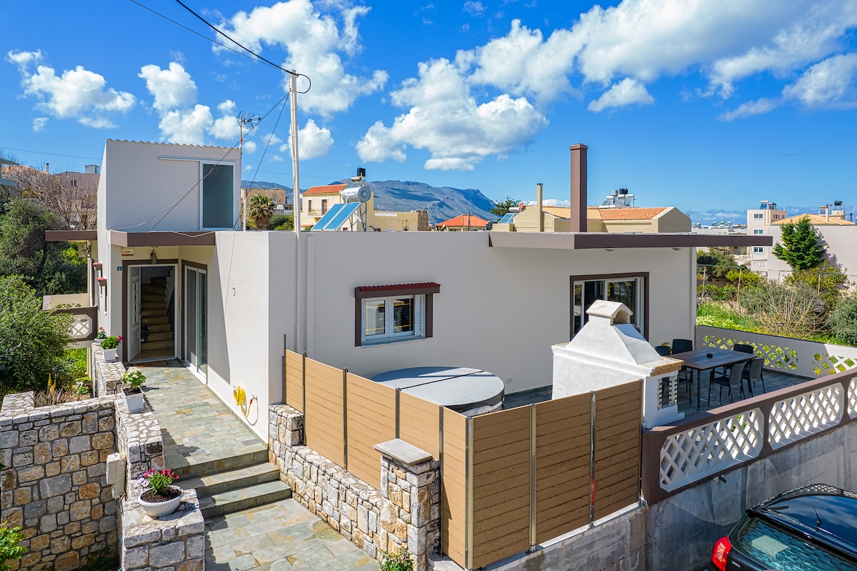 A modern house is depicted with a light-colored exterior, surrounded by greenery. A circular hot tub is visible on a patio area fenced for privacy. Steps lead to the entrance, and a dining table is seen nearby. The sky is clear with scattered clouds.
