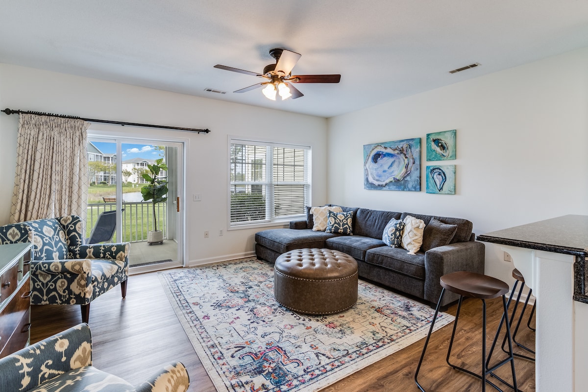 The living room features a comfortable sectional sofa and an upholstered ottoman on a decorative area rug. Windows allow natural light to filter in, while a ceiling fan provides comfort. A sliding door leads to an outdoor space with views of the surrounding community.