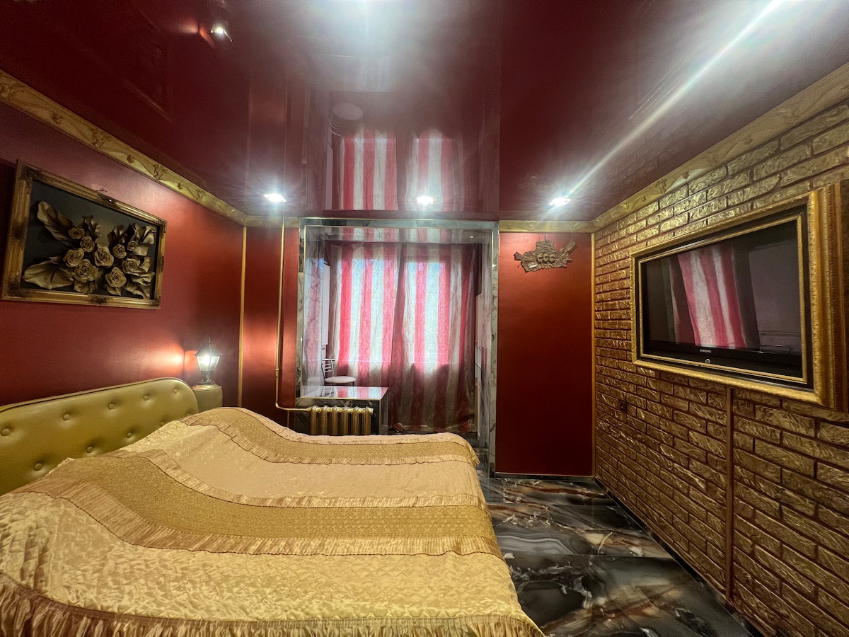 A room features rich red walls and a textured gold headboard. A spacious bed is covered with a light gold bedspread, and soft lighting illuminates the area. A window dressed with patterned curtains allows natural light to filter in, and a wall-mounted television is visible.