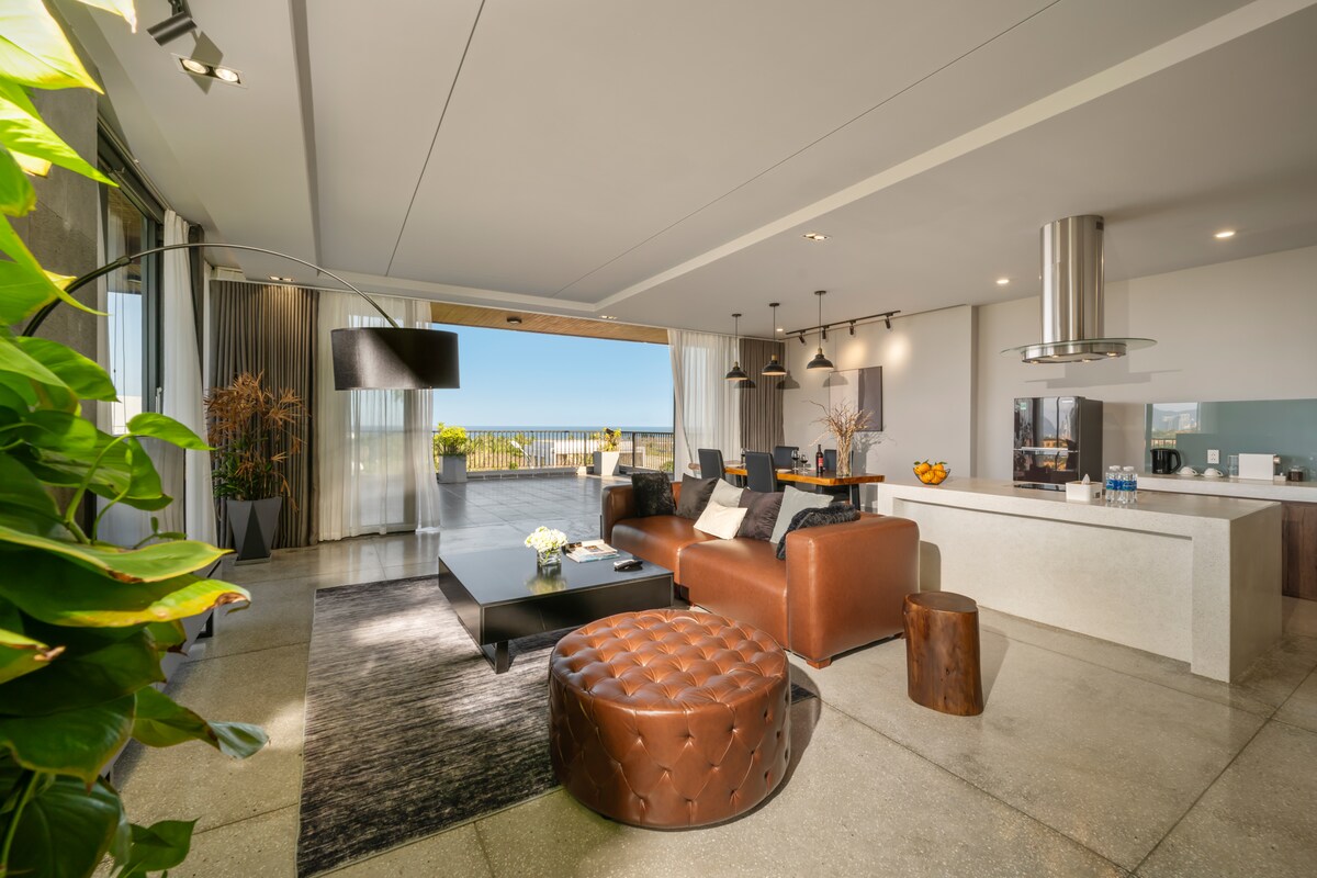 The spacious living room offers panoramic sea views visible through large bay windows, which open onto a terrace. A modern brown leather sofa and a round ottoman complement a sleek coffee table, surrounded by chairs. A well-equipped kitchen area features a stylish island, enhancing the open layout.