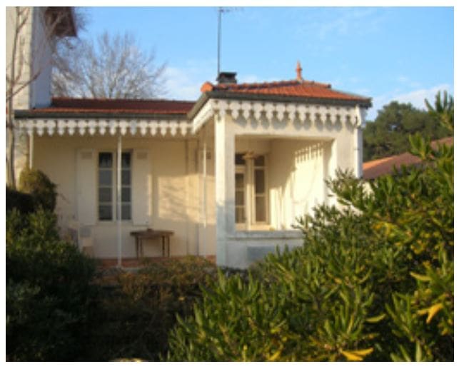 The exterior of a historic house is featured, showcasing traditional architectural elements with decorative eaves. Surrounding greenery frames the property, while a small table can be seen on the covered porch area, creating an inviting space to relax outdoors.