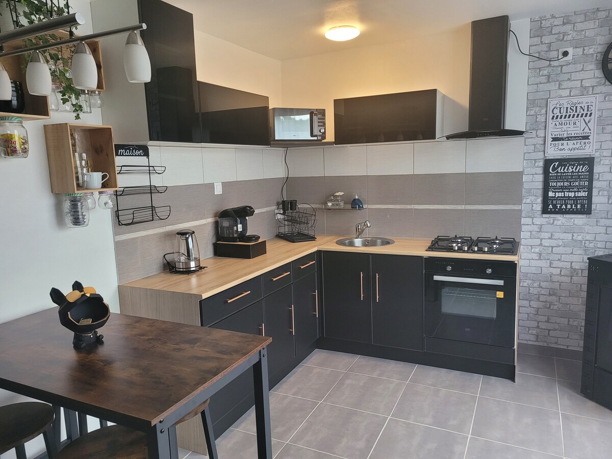 A modern kitchen is presented with dark cabinetry and light wood accents. Essential appliances include a refrigerator, microwave, coffee maker, and gas stove. A compact dining table is set for two, while the counter features a sink and various utensils, blending function and style.