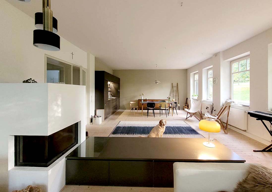 An open living space is highlighted with a modern fireplace and a cozy seating area. Large windows allow natural light to fill the room, revealing a dining area with a table and chairs. A warm glow is emitted from a floor lamp, enhancing the serene ambiance.