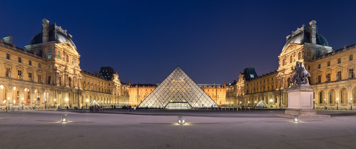 The iconic glass pyramid of the Louvre is surrounded by historic architecture, with softly illuminated facades reflecting the evening light. Statues and sculptures are positioned throughout the open courtyard, creating a harmonious blend of modern and classic design elements.