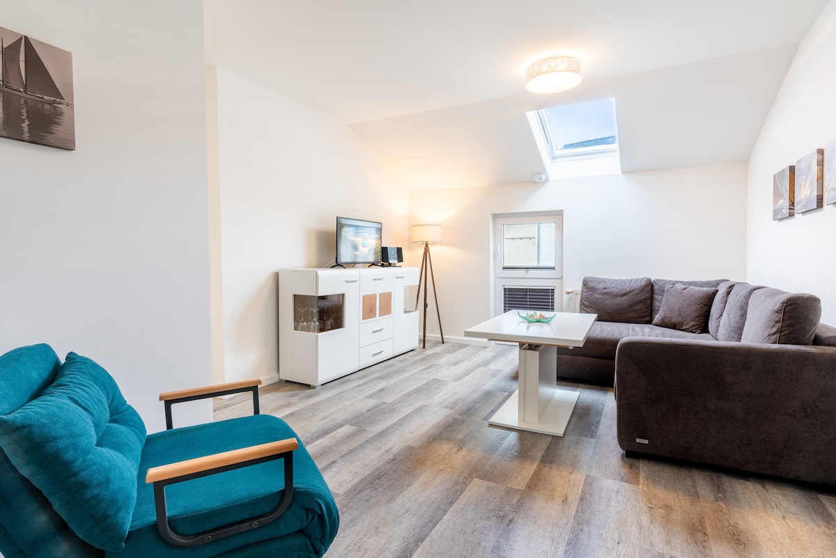 A bright living area is highlighted by natural light streaming through a skylight. A comfortable dark couch and a cyan chair face a modern white coffee table. A media unit and a floor lamp add to the space's functionality and style.