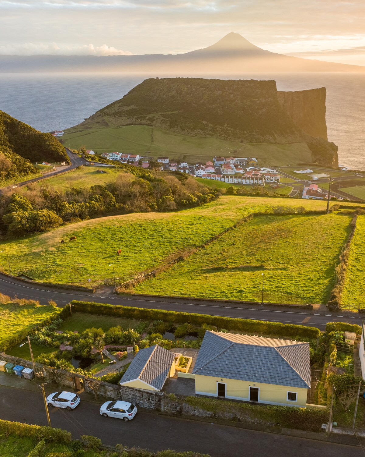 An aerial view captures a coastal landscape, featuring lush green fields and a small village nestled below. A prominent volcanic mountain looms in the distance, with the ocean stretching out to the horizon. White houses dot the scenery, complemented by winding roads.