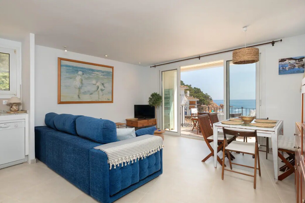 A bright living-dining area features a blue sofa, a wooden dining table with chairs, and a flat-screen TV. Large glass doors provide direct access to a terrace with views of the sea, enhancing the open and airy feel of the space.