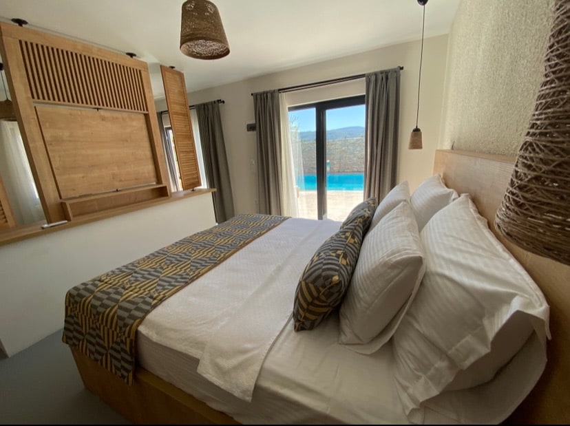 A spacious bedroom is furnished with a king size bed, complemented by decorative pillows. Large glass doors open to reveal a view of the pool area. Natural fibers in the decor enhance the room's light atmosphere, while curtains frame the windows.