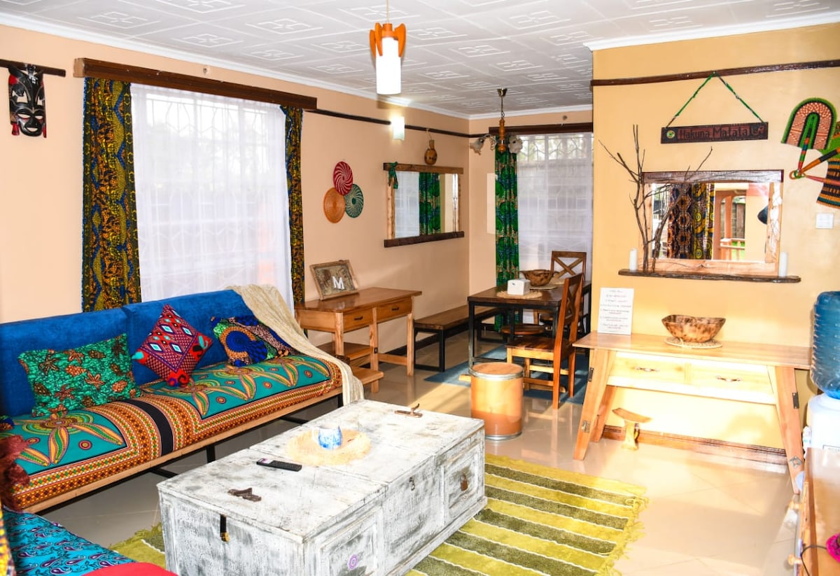 A vibrant living area features a colorful textile sofa adorned with cushions, a rustic coffee table, and a wooden dining table. Natural light brightens the space through large windows, framed by patterned curtains. Decorative artifacts on the walls add cultural charm.