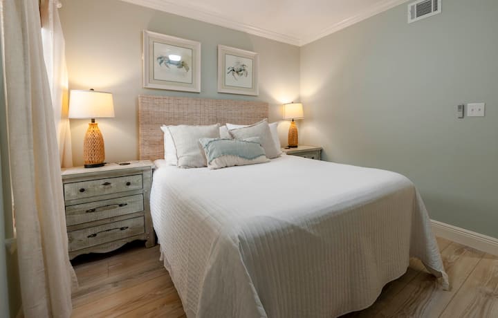 Unwind in a serene bedroom at “Gone Coastal” in Gulf Shores, AL, with a wicker headboard, queen bed, soft lighting, and elegant decor.
