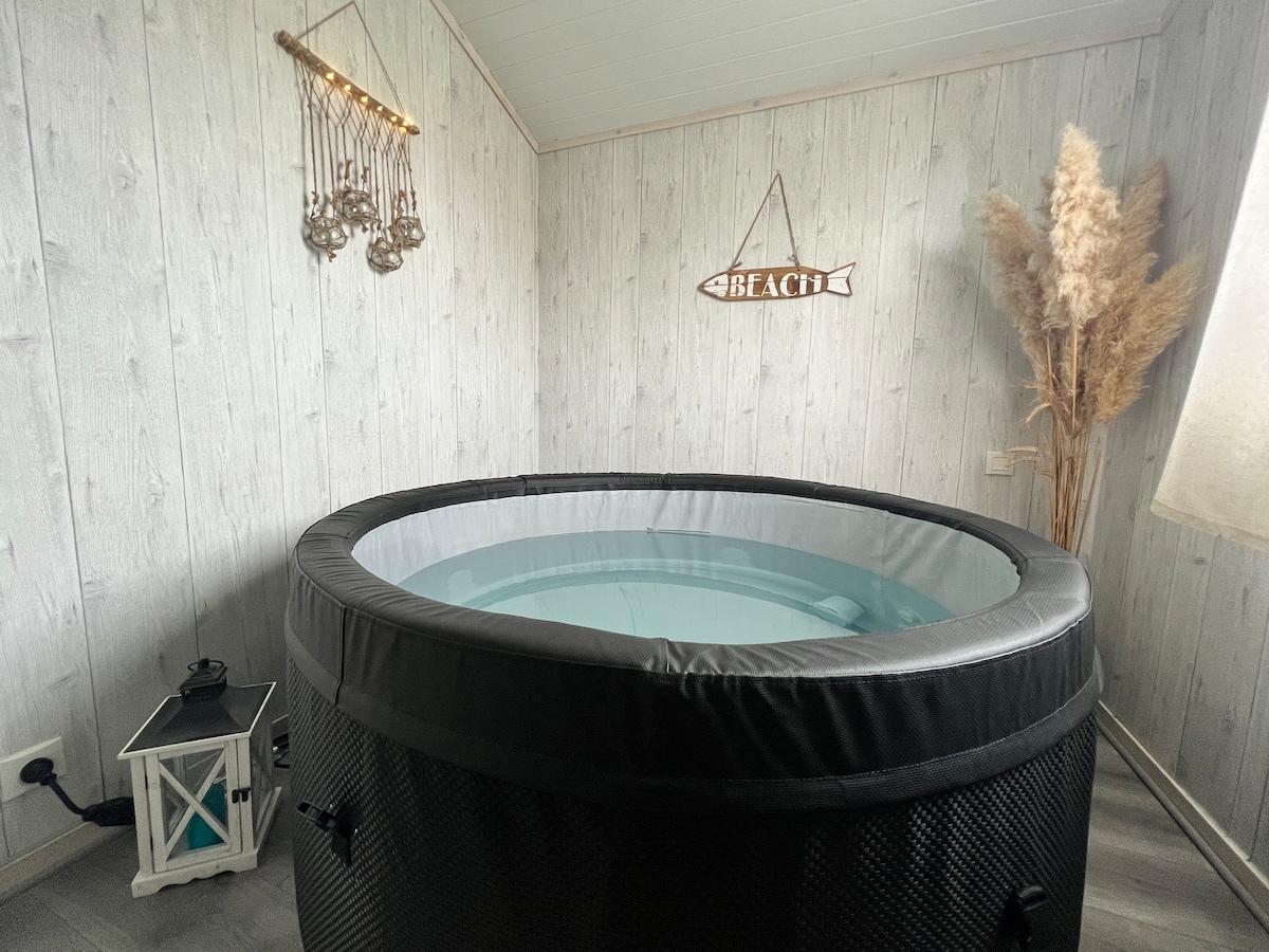 An inviting hot tub is centrally located within the room. Soft lighting from a nearby lantern creates a calm atmosphere. Decor elements include a decorative sign in the shape of a fish and dried grass arrangements, adding a natural touch to the space.