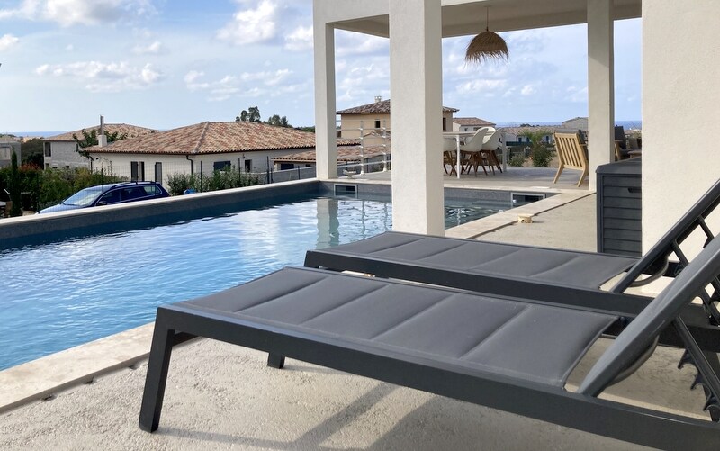 A serene pool area features a long, rectangular pool surrounded by smooth stone decking. Two lounge chairs positioned beside the pool provide a place to relax. The view beyond includes distant rooftops and a clear sky with scattered clouds.