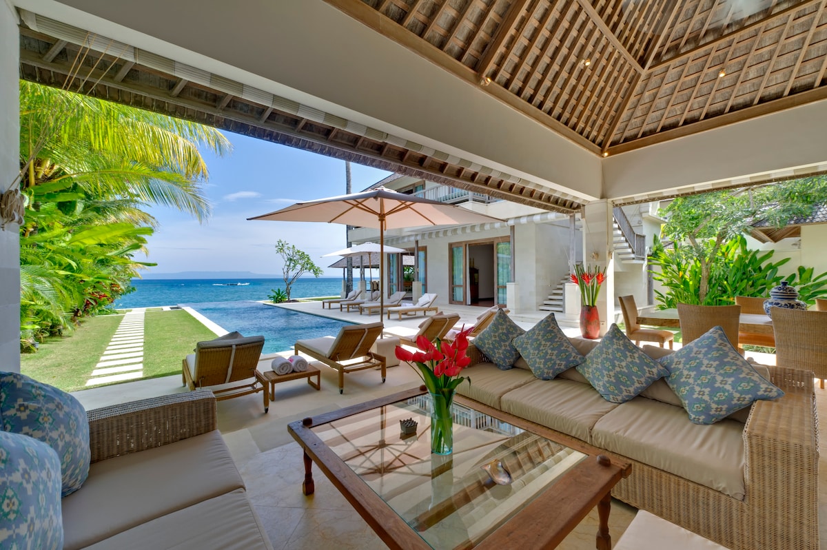 An airy alfresco lounge features a comfortable sectional sofa adorned with patterned cushions. A glass coffee table sits in front. Beyond, a clear view of the pool and ocean is visible, accompanied by sun loungers under large umbrellas. Lush greenery frames the scene.