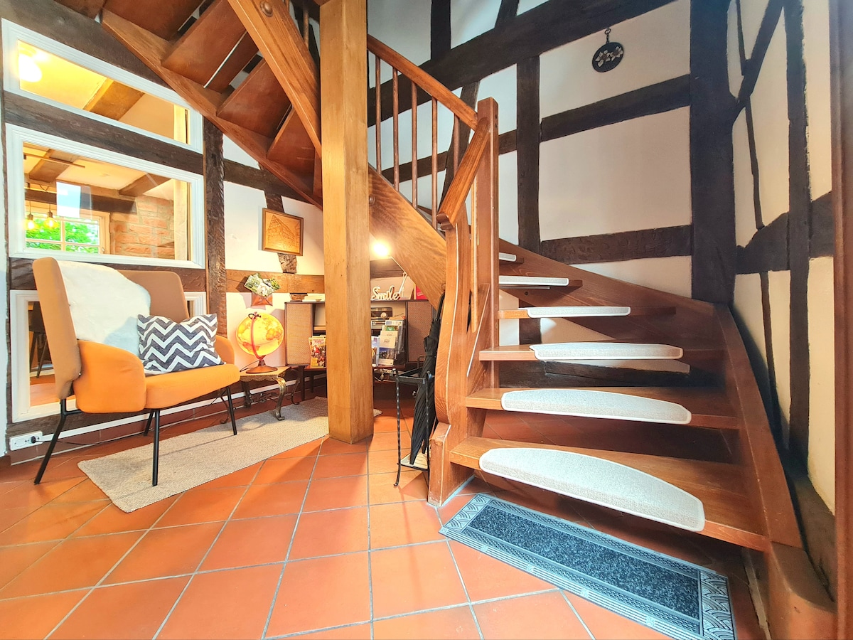 A wooden staircase with a modern design leads to the upper levels, framed by characteristic timber-framed walls. A small seating area is positioned nearby, featuring a chair with a geometric pattern and a floor lamp. Natural light enters from a window, highlighting the warm tones of the space.