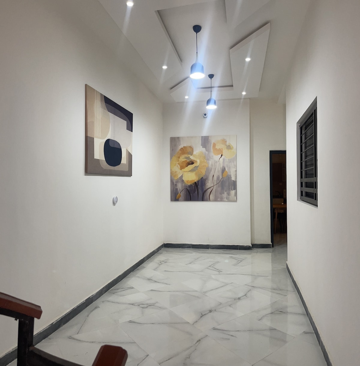 A well-lit hallway is lined with two pieces of artwork, featuring abstract shapes and floral designs. The ceiling includes modern light fixtures, while polished marble flooring adds a touch of elegance to the space.