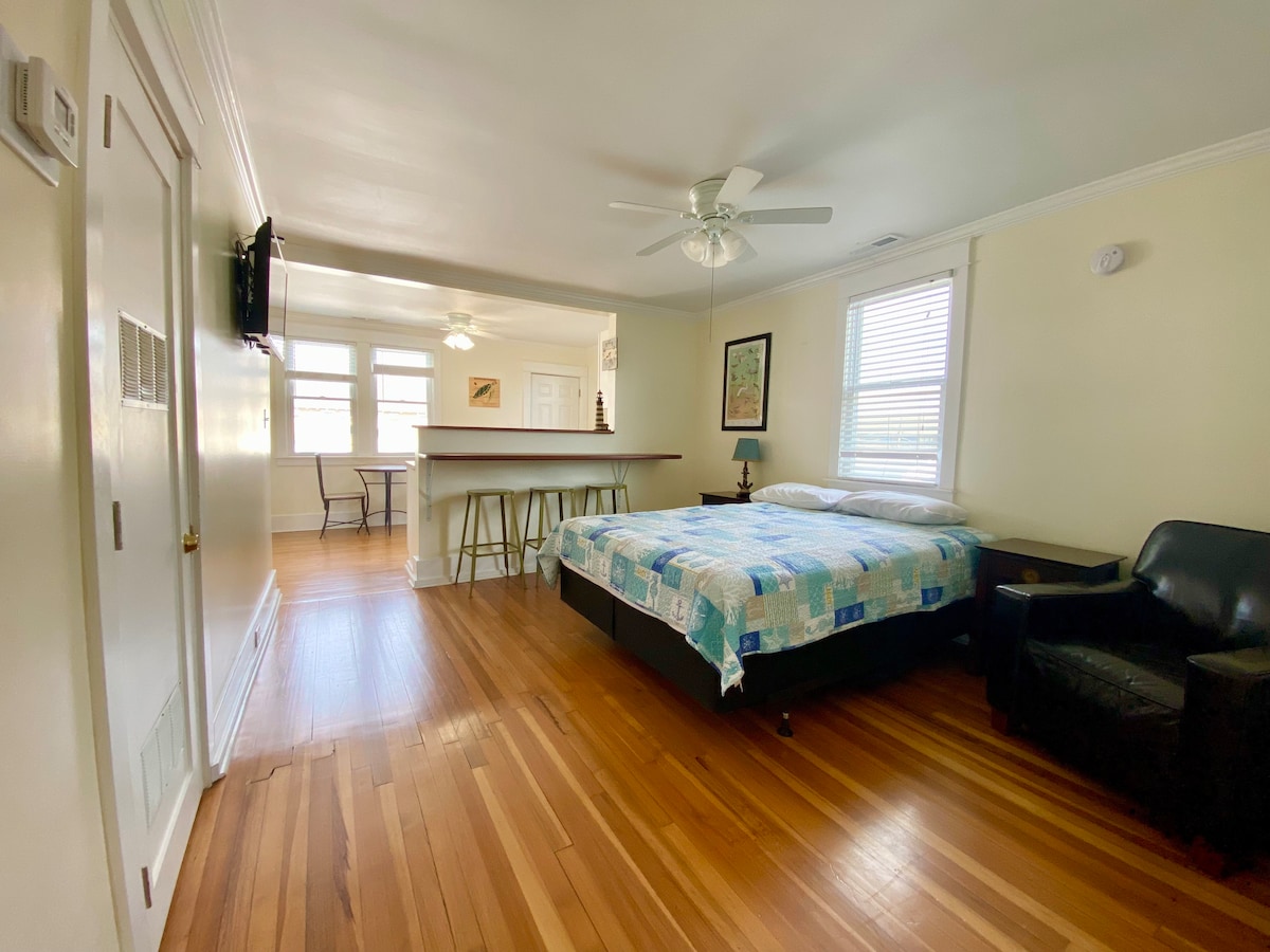 An efficiency apartment features a spacious bed covered with a patterned quilt. A comfortable armchair is positioned nearby, and light hardwood flooring enhances the openness of the layout. In the background, a bar area with stools is visible, contributing to a welcoming living space.