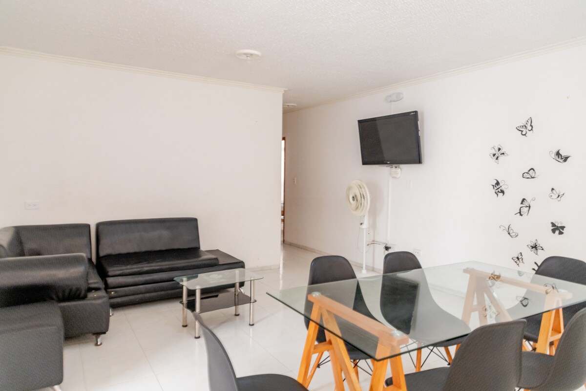The living area features a black sectional sofa and a glass-top coffee table, creating a functional seating space. A modern dining table with wooden legs is surrounded by dark chairs. A wall-mounted television and decorative wall art of butterflies add to the decor.