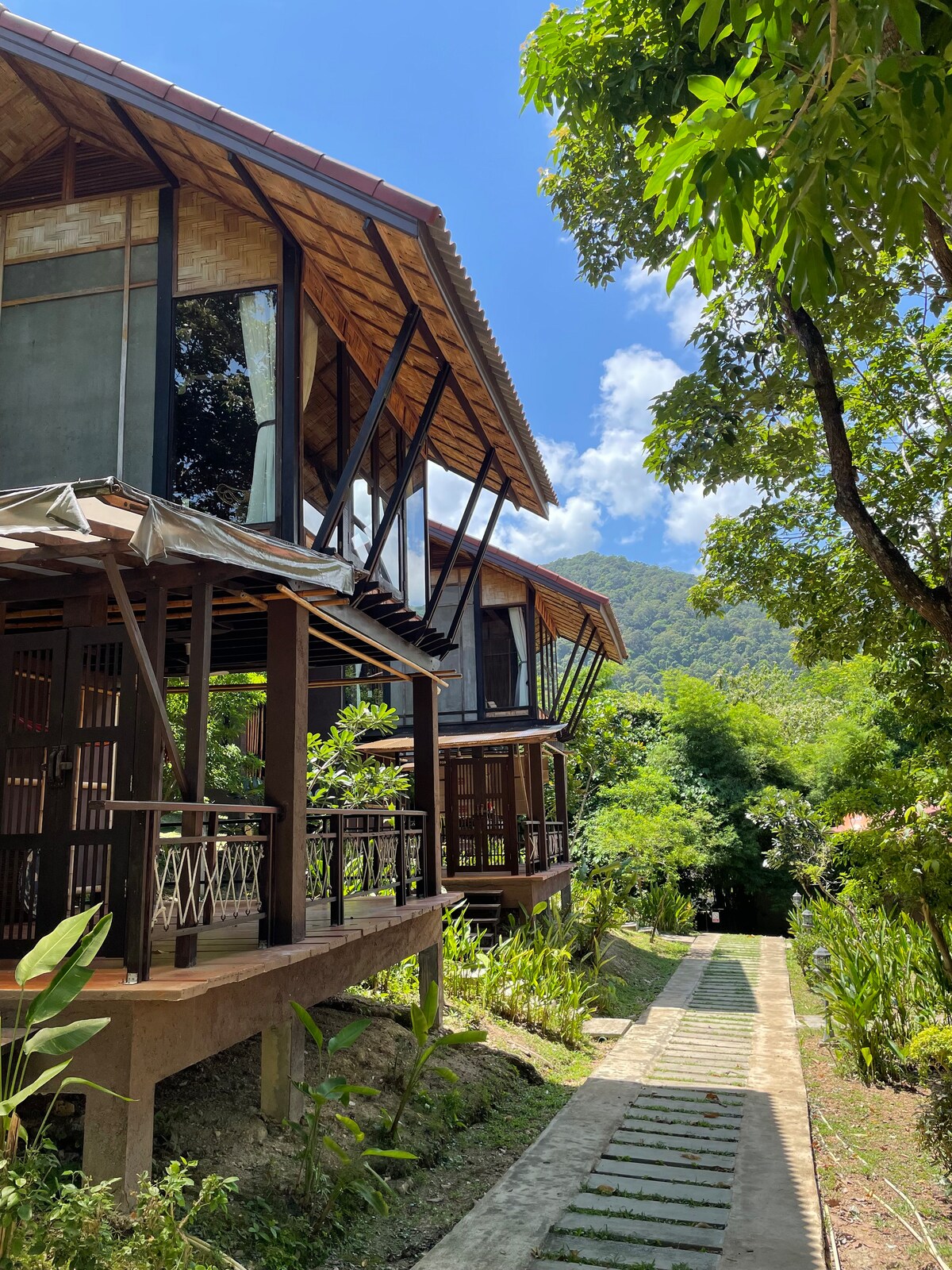 Two elegantly designed villas feature large windows and wooden structures, set amidst lush green foliage. A stone pathway leads through the garden, connecting the buildings to the surrounding landscape. Clear blue skies and scattered clouds enhance the inviting atmosphere of the resort.