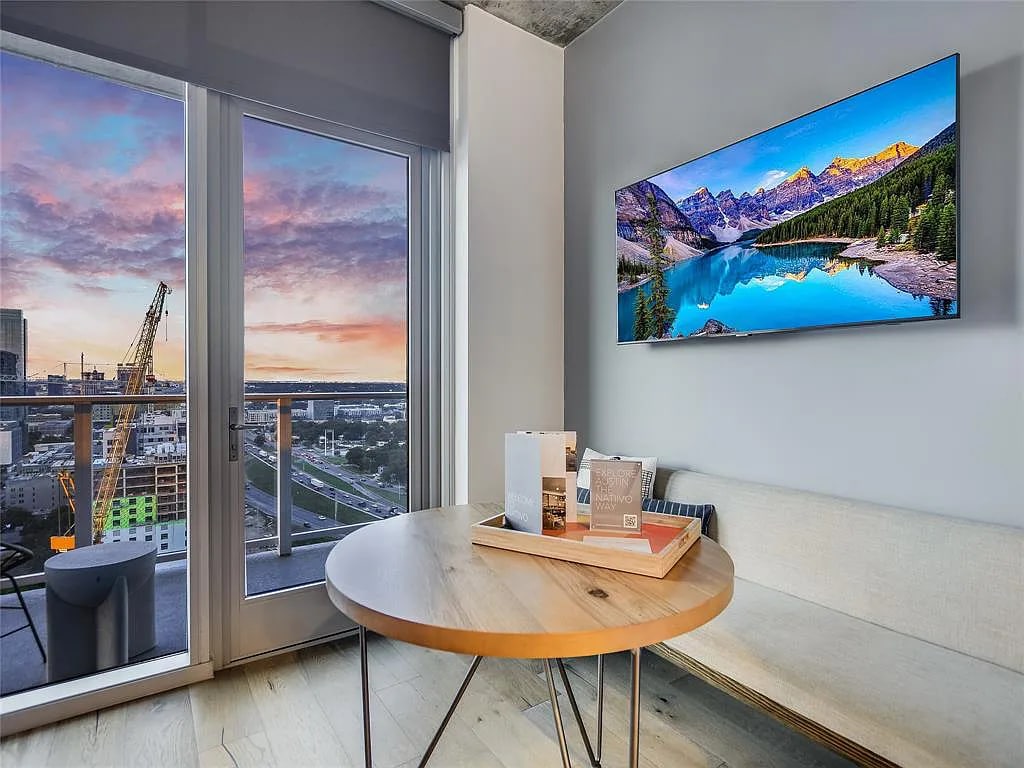 A round wooden table is positioned beside a large window, offering a view of a colorful sunset. On the table, a neutral-colored decorative tray holds several items. A flat-screen TV displays a serene landscape featuring mountains and water.