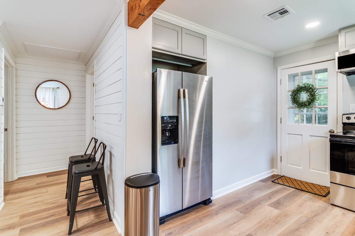 A modern kitchen is shown, featuring stainless steel appliances, including a refrigerator and an oven. A wooden door with a glass panel opens to the outside, adorned with a green wreath. Bar stools are positioned against the wall, and natural light brightens the space.