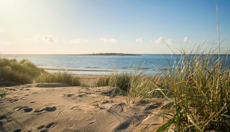 A sandy beach is captured in the foreground, with gentle dunes covered in green grass leading to a calm, clear sea. The horizon showcases a distant landmass, under a bright blue sky scattered with soft clouds.