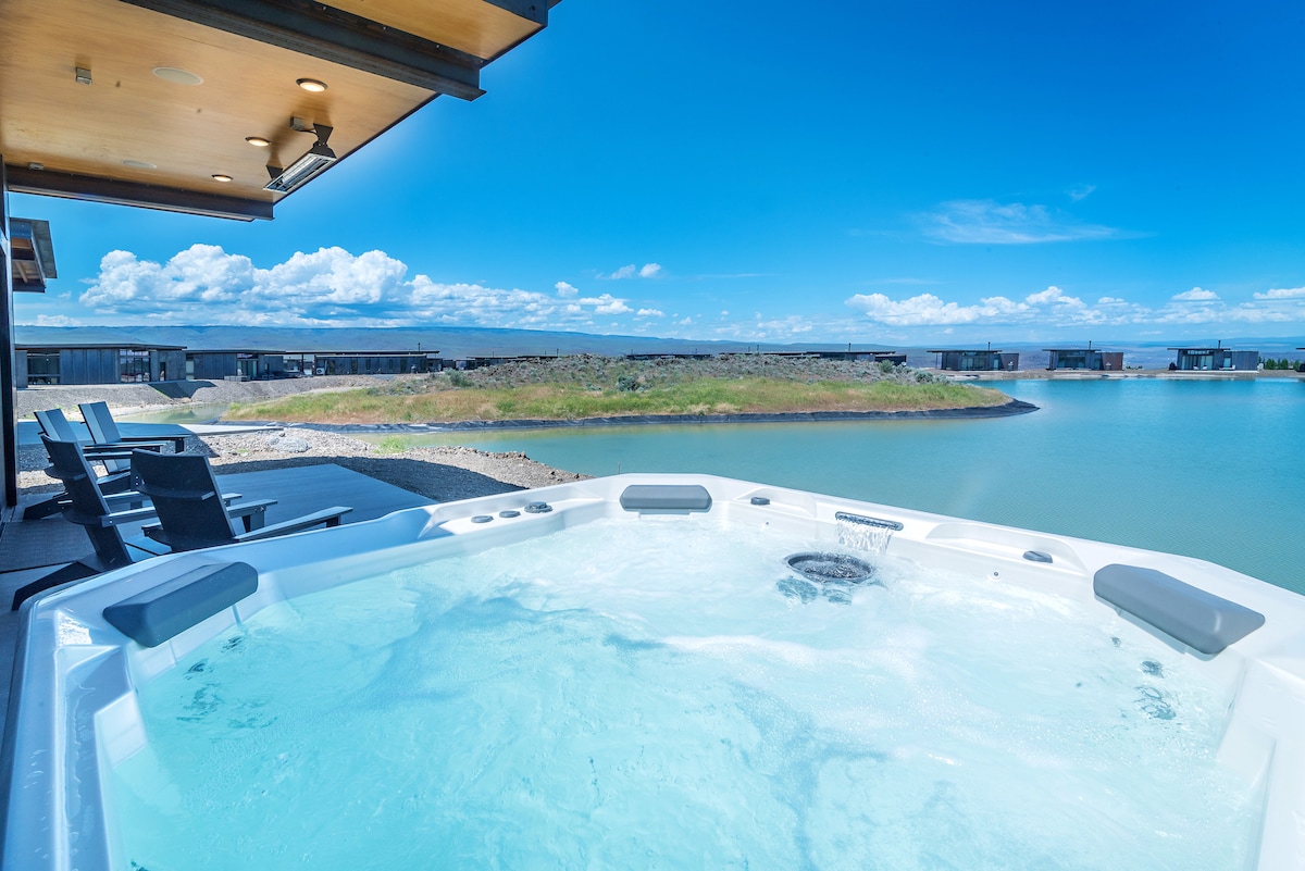 A hot tub is positioned on a patio, offering views of the surrounding lake and landscape. Sunlight reflects off the water, and a series of lounge chairs are arranged nearby. The sky is bright with a few clouds.