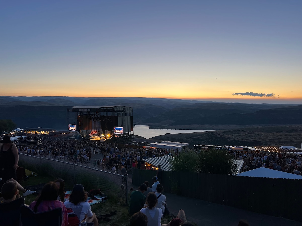 A wide view of a concert at the Gorge Amphitheatre reveals a vibrant crowd gathered below a large stage, bathed in warm sunset hues. A calm lake reflects the sky’s colors, with rolling hills in the distance enhancing the scenic backdrop.