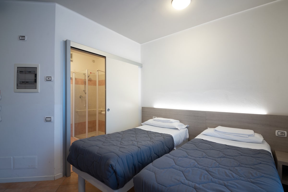 A room is presented with two single beds featuring simple wooden headboards and soft bedding. The light-colored walls complement the natural light from the window. A private bathroom is visible through a glass door, showcasing a walk-in shower.