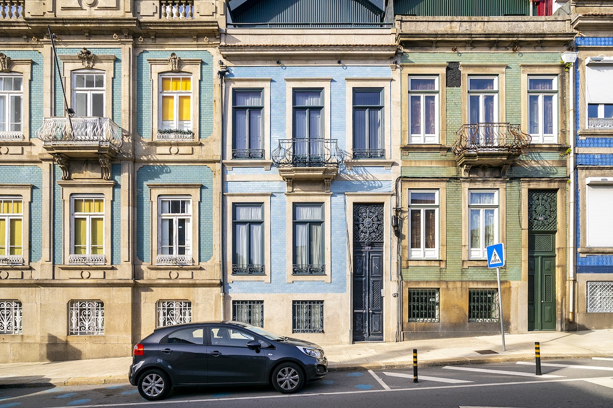 A row of colorful vintage buildings showcases architectural features typical of the XIX century, with detailed balconies and large windows adorned with shutters. A parked car is visible in the foreground, along with street signage, illustrating the urban setting.