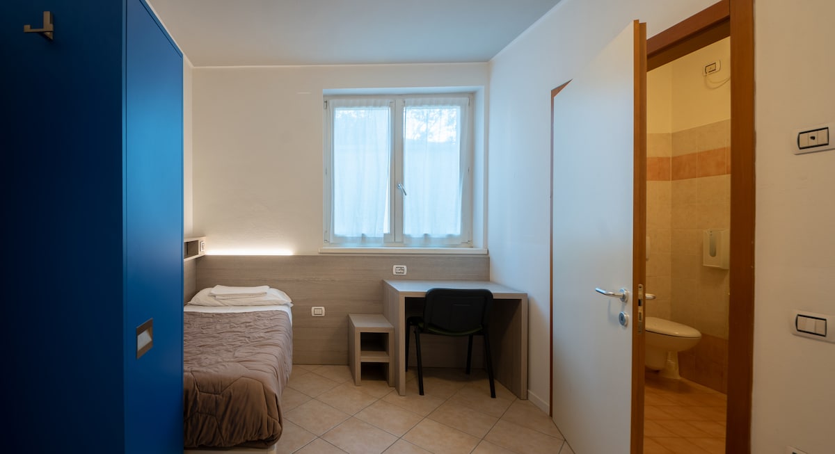A simple room with a single bed covered by a light blanket is shown. A blue wardrobe and a desk with a chair are visible. Natural light filters through a window dressed with sheer curtains, and a bathroom door is seen in the background.