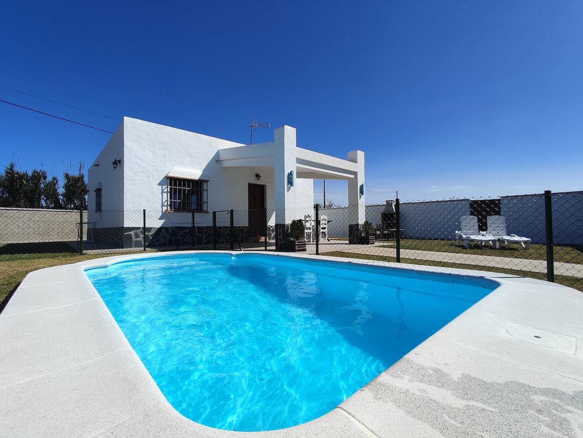 A well-maintained outdoor area features a clear blue pool surrounded by a spacious grassy lawn. A white, single-story house with a covered terrace is visible in the background, along with sun loungers and outdoor furniture. A secure fence outlines the property, enhancing privacy.
