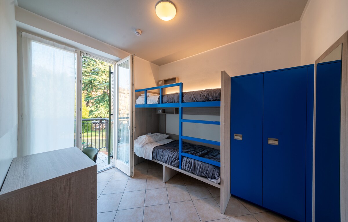 A room designed for comfort features a double bunk bed, complemented by a simple wooden wardrobe. Natural light floods in from a sliding glass door, which opens to a balcony overlooking the garden. The room's neutral tones create a calming atmosphere.
